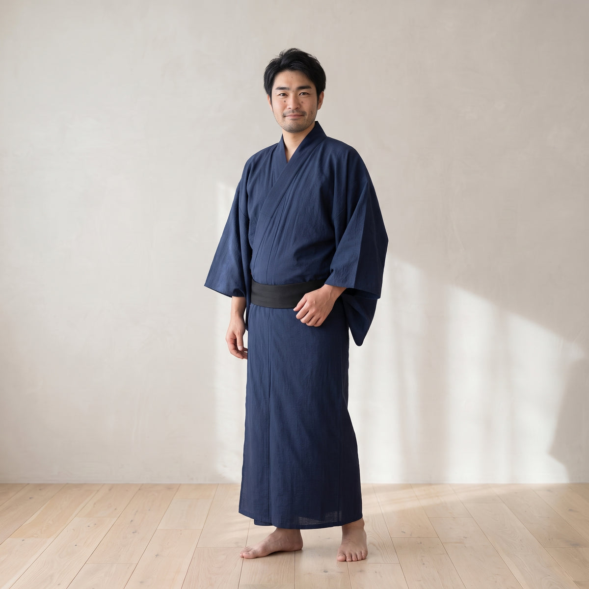 Homme portant le yukata japonais bleu marine uni serré par une ceinture noire, debout pieds nus dans une pièce claire